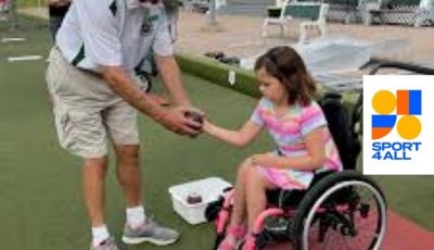 a young girl in a wheelchair being handed a bowling ball from a bowling coach on the green