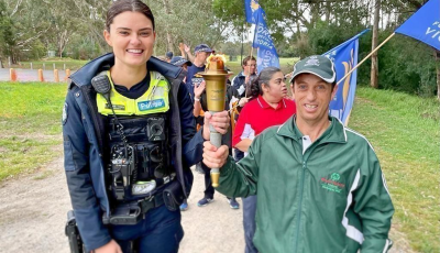 A woman wearing a police vest and a man wearing a green tracksuit both hold an Olympic torch and are smiling at the camera