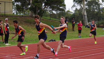 On an athletics track, an athlete wearing a club vest is handing over a baton to an athlete getting ready to run.