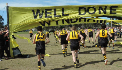 a group of men wearing yellow and black sports clothing run under a yellow banner saying Well Done 