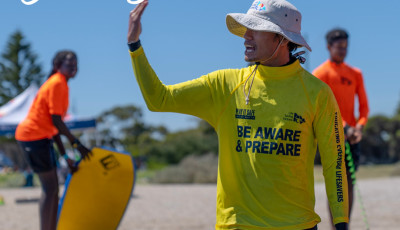 A man in a yellow top and white hat is beckoning away from the camera. Two children in orange tops are further behind on the beach.