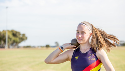 a girl wearing an athletics vest is holding a shotput ready to throw