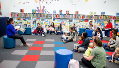 Parents and children sitting in the library listening to a librarian read a story.