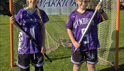 a boy and girl in purple and black sports uniforms stand infront of a goal holding Lacrosse sticks