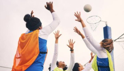 a group of girls wearing netball bibs are reaching up to a netball in the air