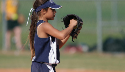 a girl is holdng up a baseball glove and ball, ready to bowl