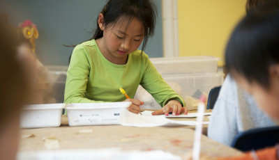 A girl writing on paper in a bright room with other children.